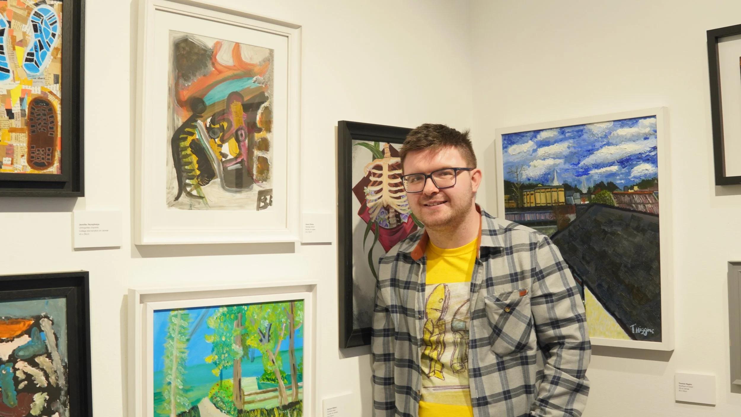A person stands in an art gallery in front of several framed abstract paintings and a painting of a cityscape.