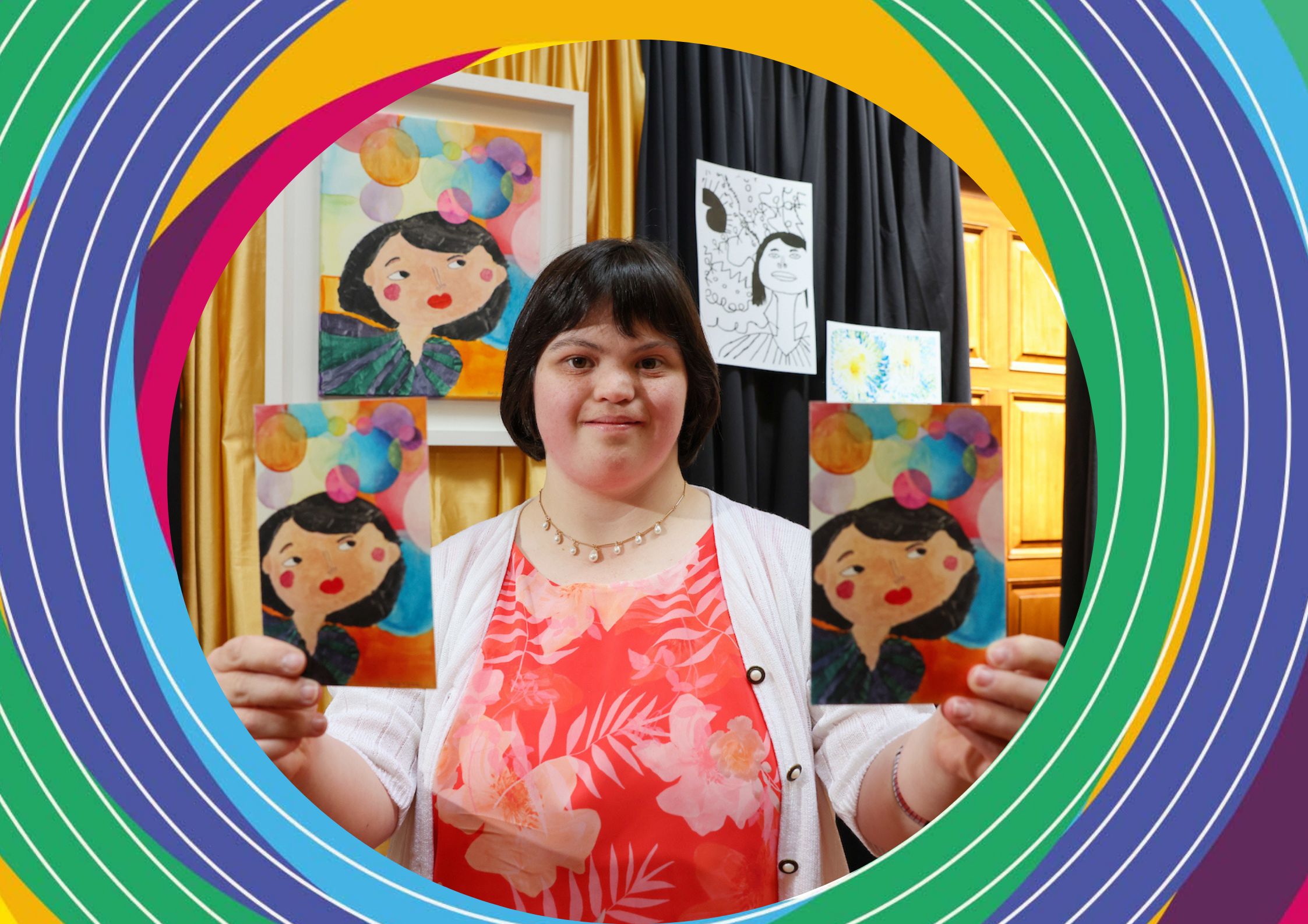Artist Amy Clarke, a woman wearing a floral red top and a white jacket, holds two prints of her artwork of a woman on a colourful circle background.