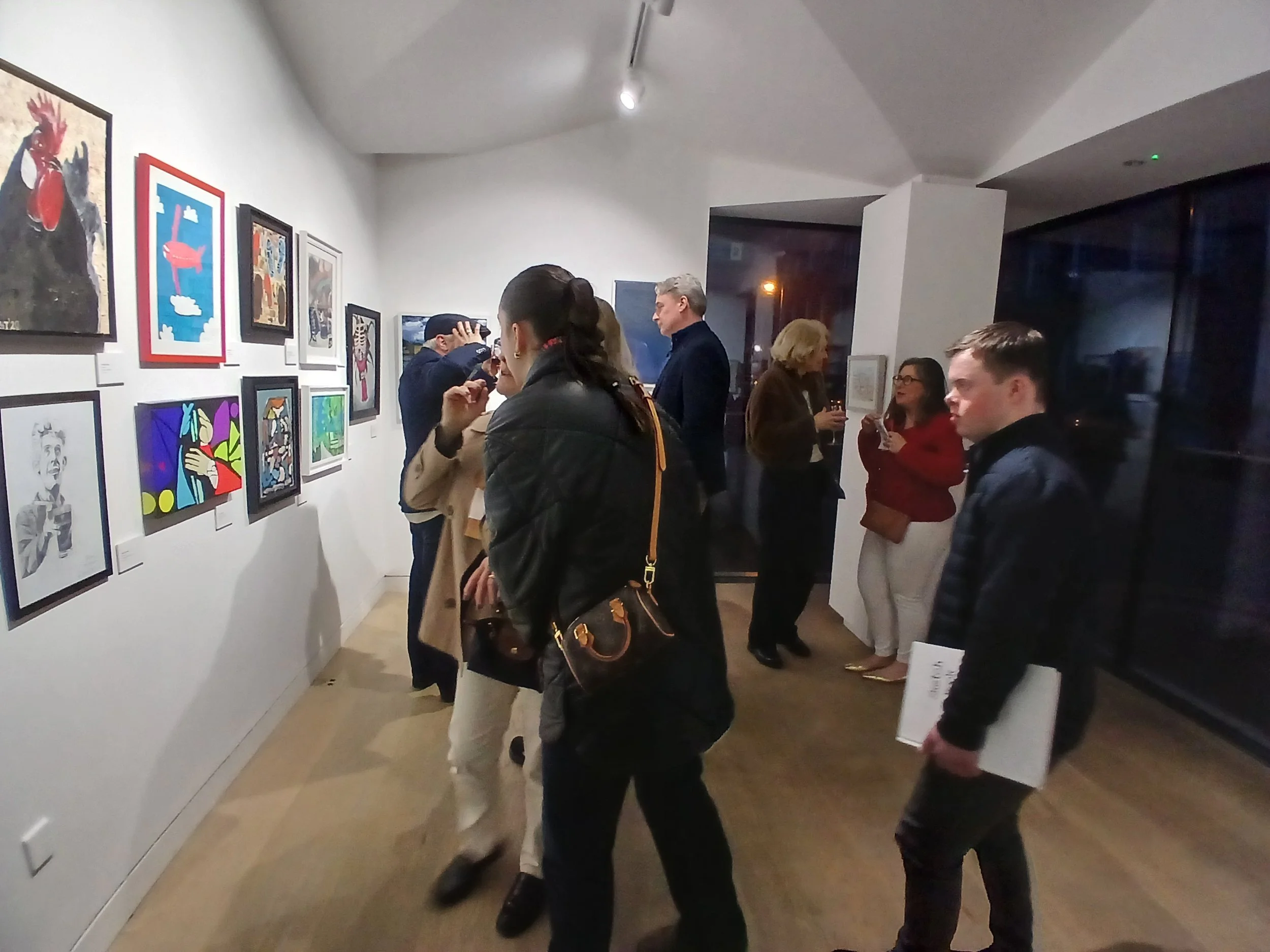 Attendees observe colorful and varied artwork displayed on white walls in a gallery exhibition.