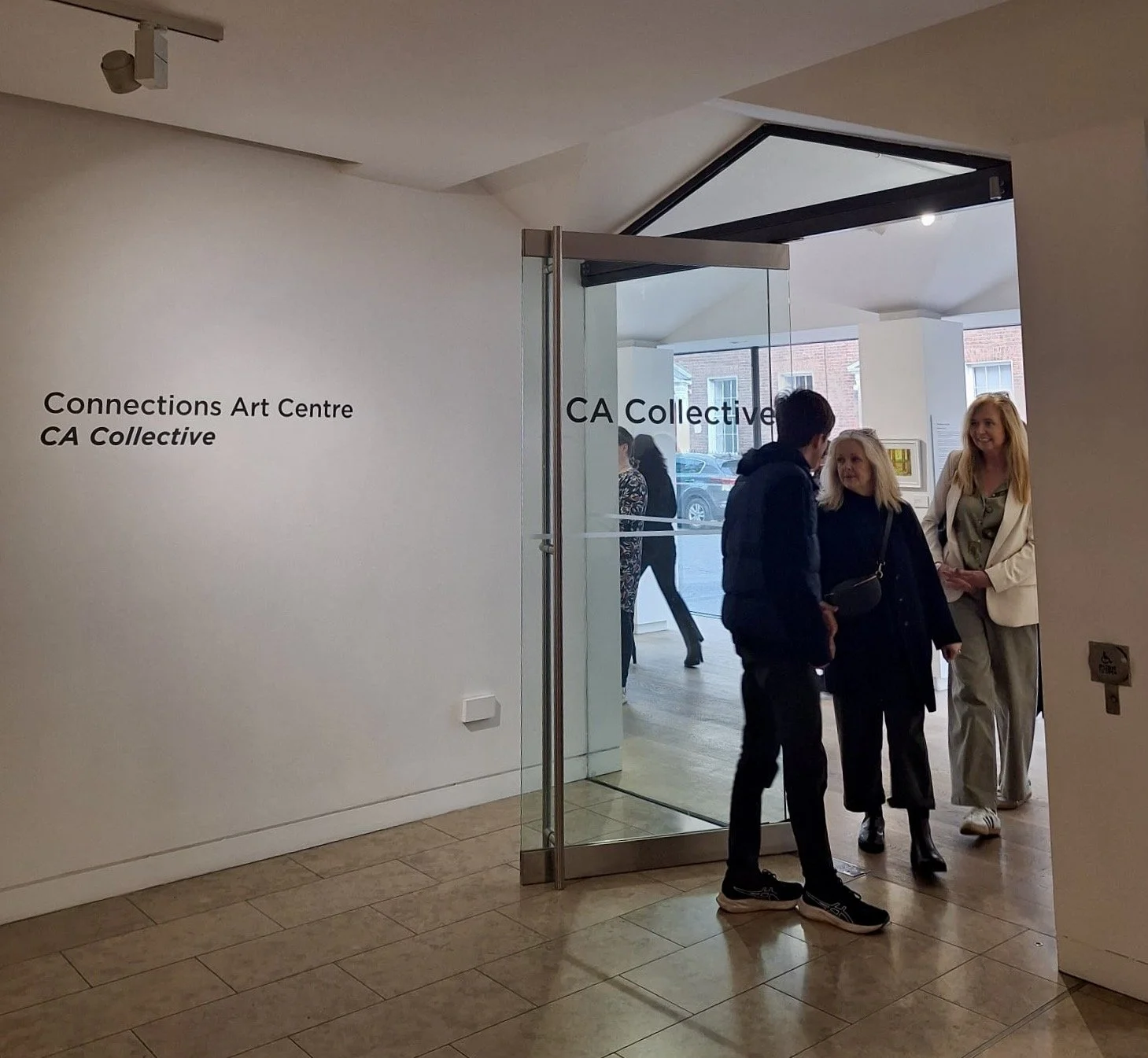 People entering a contemporary art gallery featuring "Connections Art Centre CA Collective" signage on a white wall and frosted glass doors.