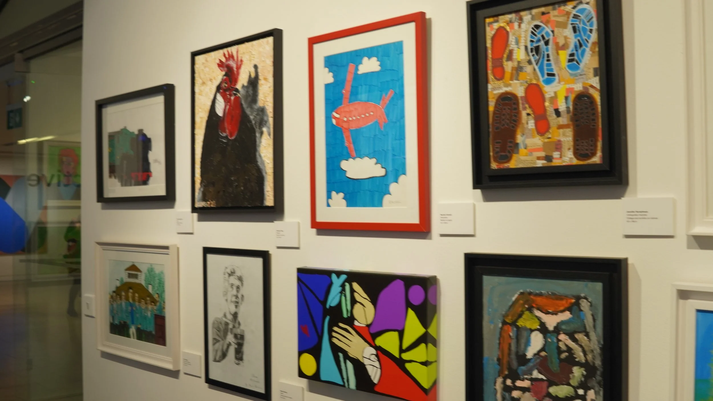 Wall of framed artwork in an exhibition, including a rooster, a plane in the sky, and shoe prints.