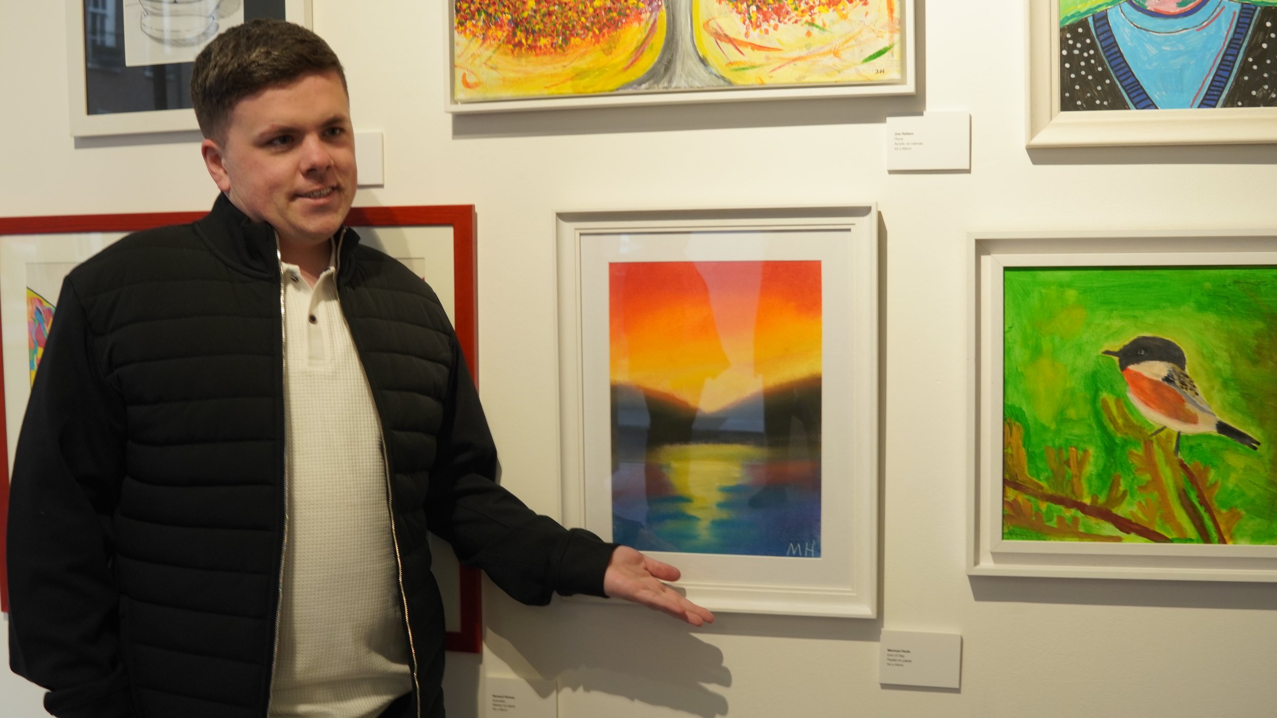 A man in a black puffer jacket and white polo shirt stands in front of a gallery wall featuring several framed artworks, gesturing towards a framed painting of a colorful sunset reflected in water.