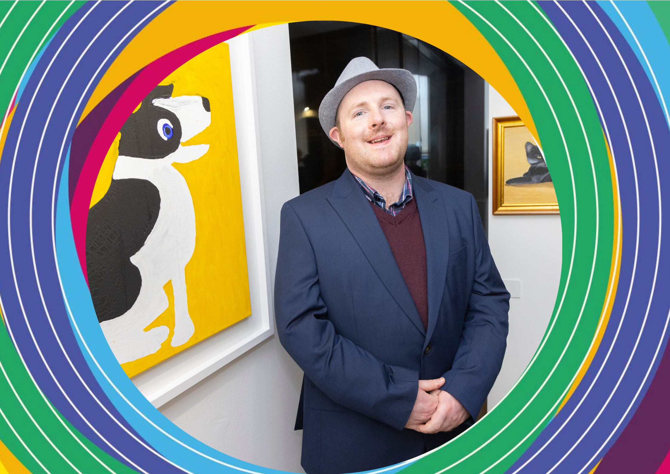 Artist Matthew Sexton, a man wearing a navy suit and a grey fedora stands beside his artwork of a dog on a yellow background.