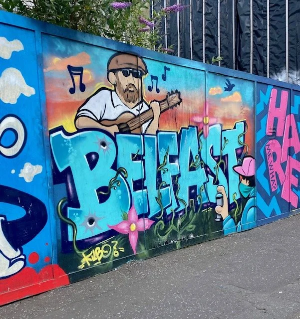 Graffiti mural featuring a cartoon man playing a guitar with musical notes, the word "BELFAST" in large blue lettering, and a figure spraying graffiti, all against a sunset sky background with clouds and a bird.