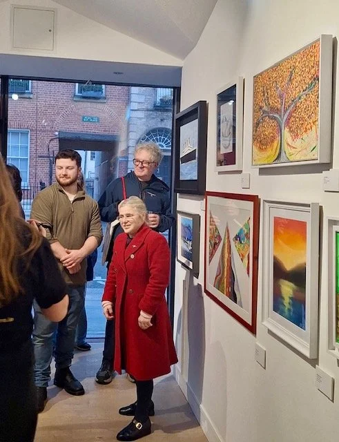 A group of people stand in a well-lit art gallery, looking at various framed paintings on a white wall. The paintings include a colorful abstract tree, a landscape with a sunset over water, and a piece with geometric shapes. The viewers are casually 