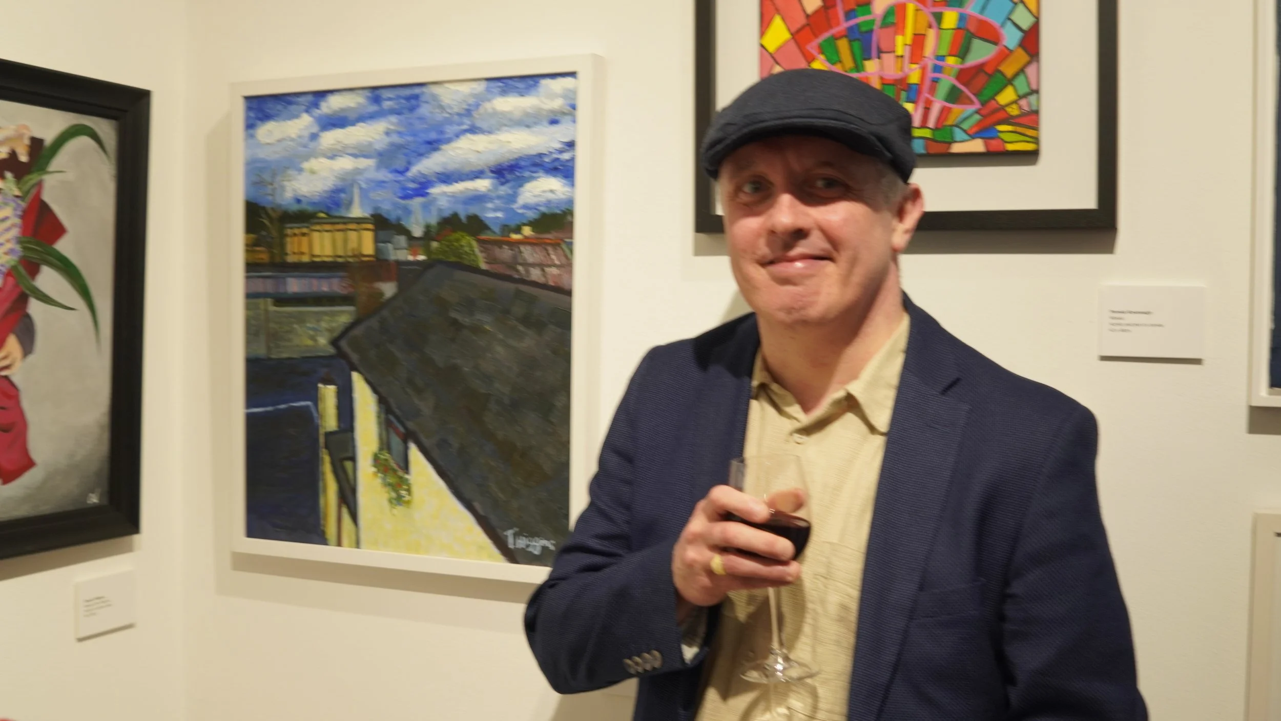 A man in a navy blue blazer and a flat cap stands in an art gallery, holding a glass of red wine, with colorful paintings on display behind him.