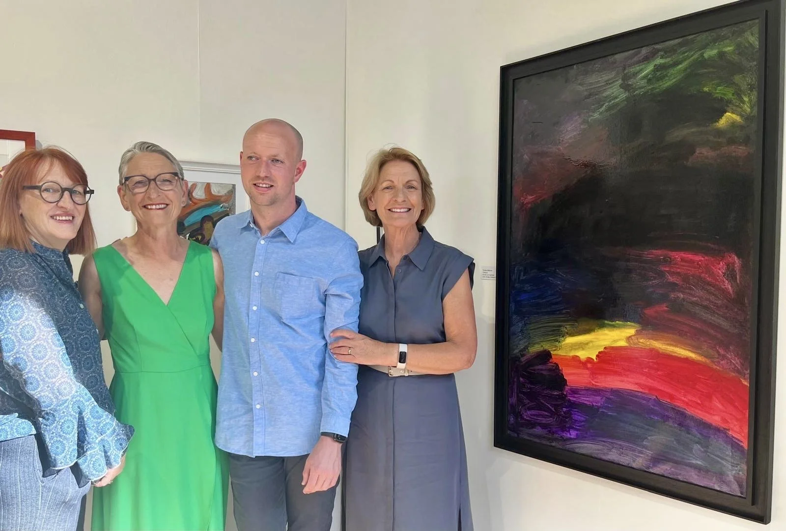 Four people stand smiling in front of a large, abstract painting with dark hues and streaks of vibrant color.