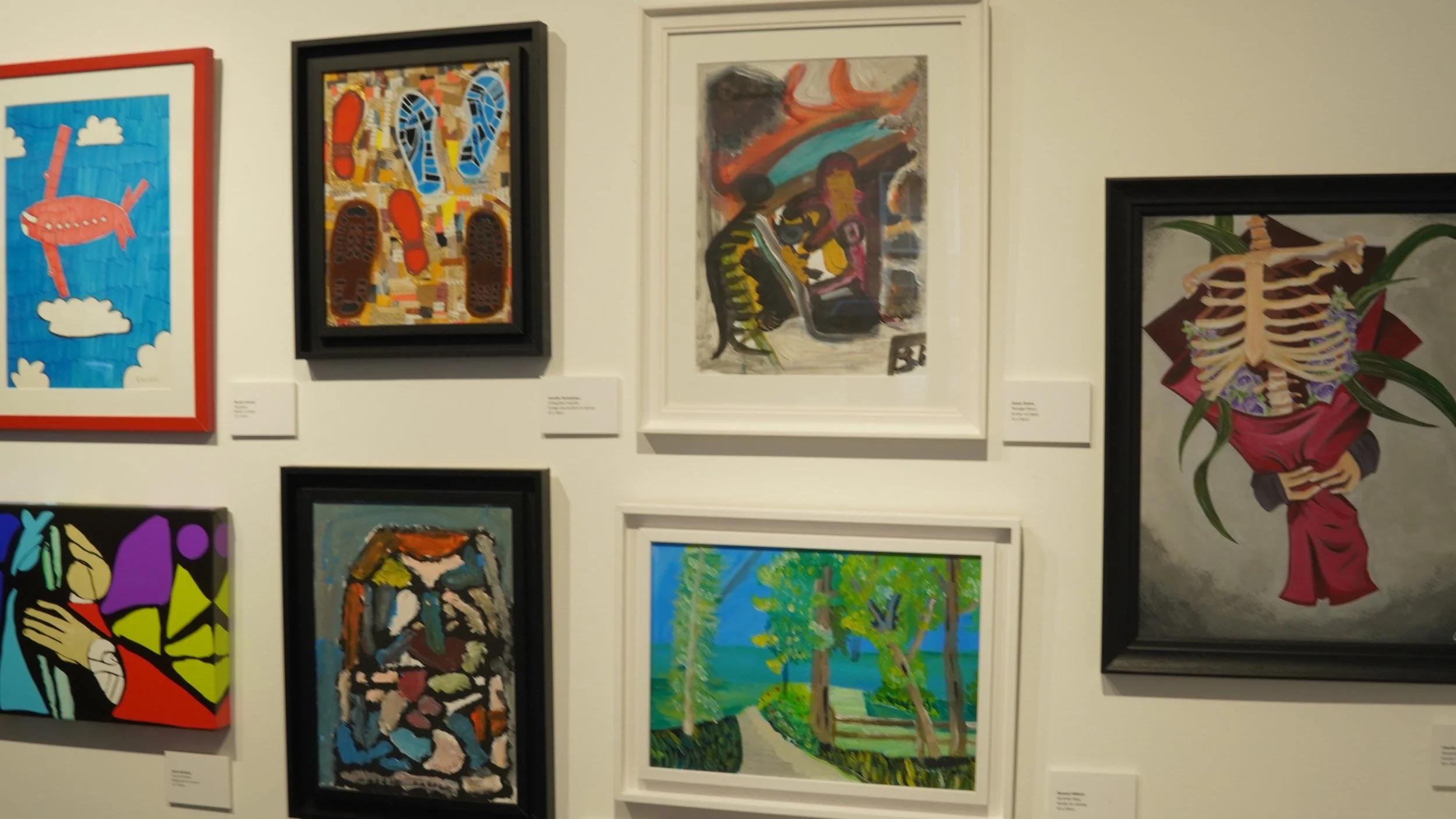Wall display of various framed artworks including a red airplane in a blue sky, shoe soles, abstract figures, a landscape with trees, and a skeleton with flowers.