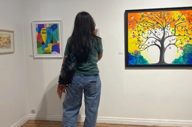 A person viewed from behind stands in an art gallery, looking at two paintings on a white wall: a colorful abstract portrait on the left and a vibrant tree of life on the right.