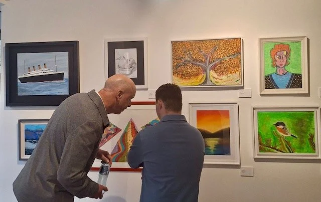 Two people examine a wall of diverse paintings in an art gallery. Paintings visible include the Titanic ship, a still life of apples, a colorful tree, a portrait, a landscape with a sunset over water, a colorful abstract piece, a mountain landscape, 