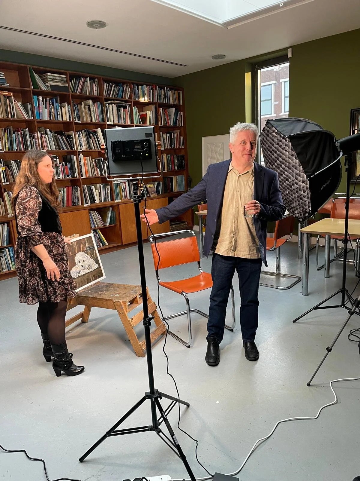 A man in a navy blue blazer and a flat cap stands in an art gallery, holding a glass of red wine, with colorful paintings on display behind him.