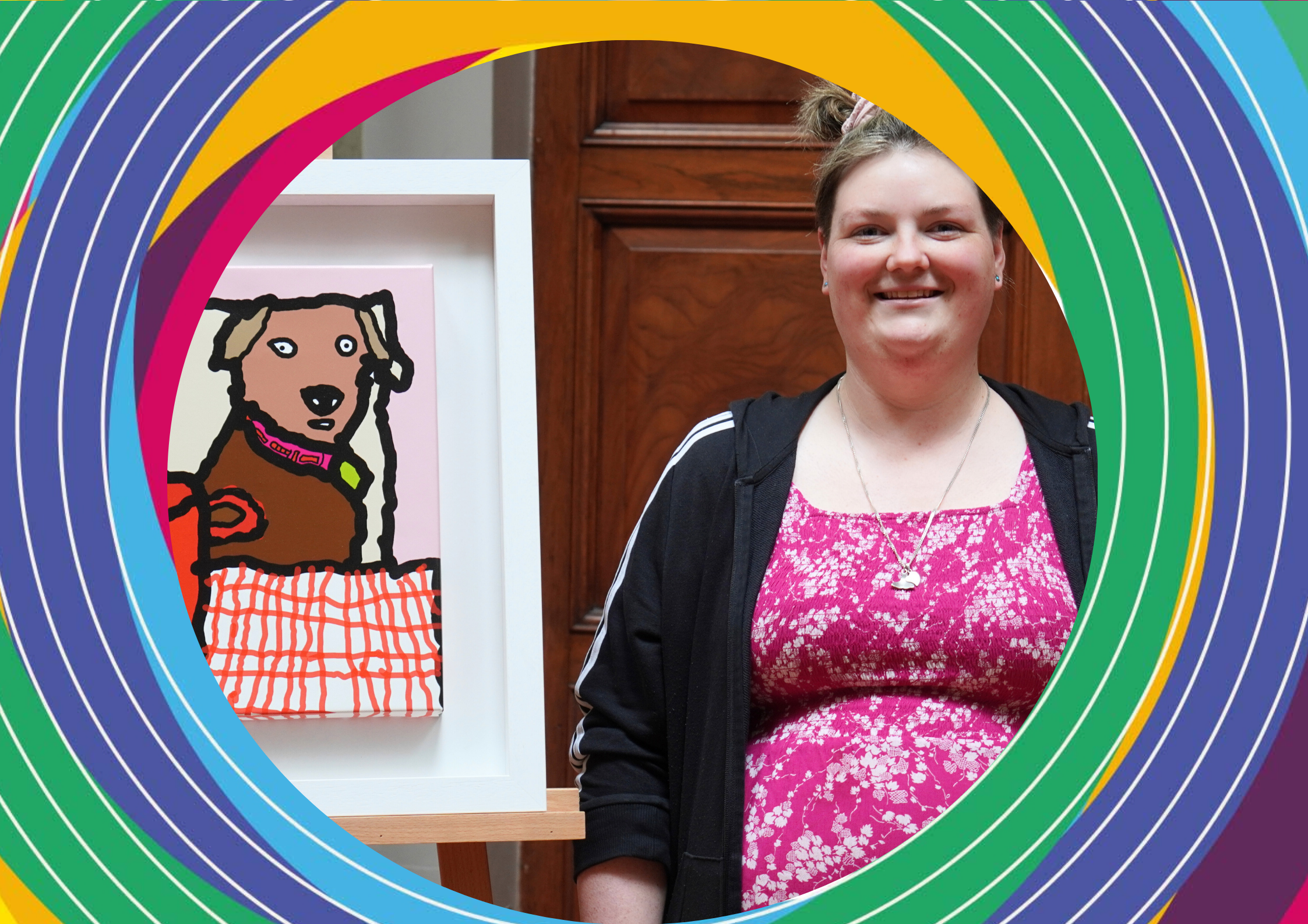 Artist Sarah McNulty, a woman wearing a pink floral top stands beside her artwork of a dog at a kitchen table with a cup of tea.