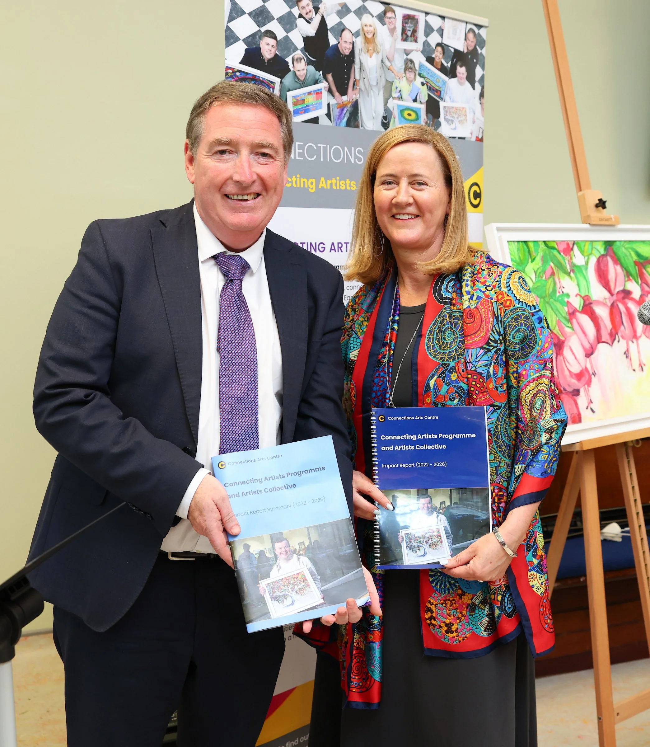 A man and woman stand smiling, holding two brochures titled "Connecting Artists Programme and Artists Collective Impact Report (2022-2026)" in front of a banner displaying artwork and text.