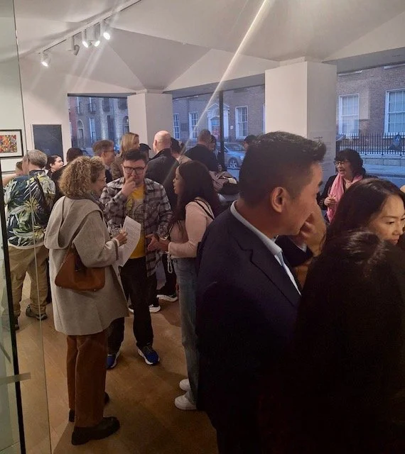 A diverse group of people mingle at an art gallery opening, with artwork visible on the walls and large windows looking out onto a street scene.