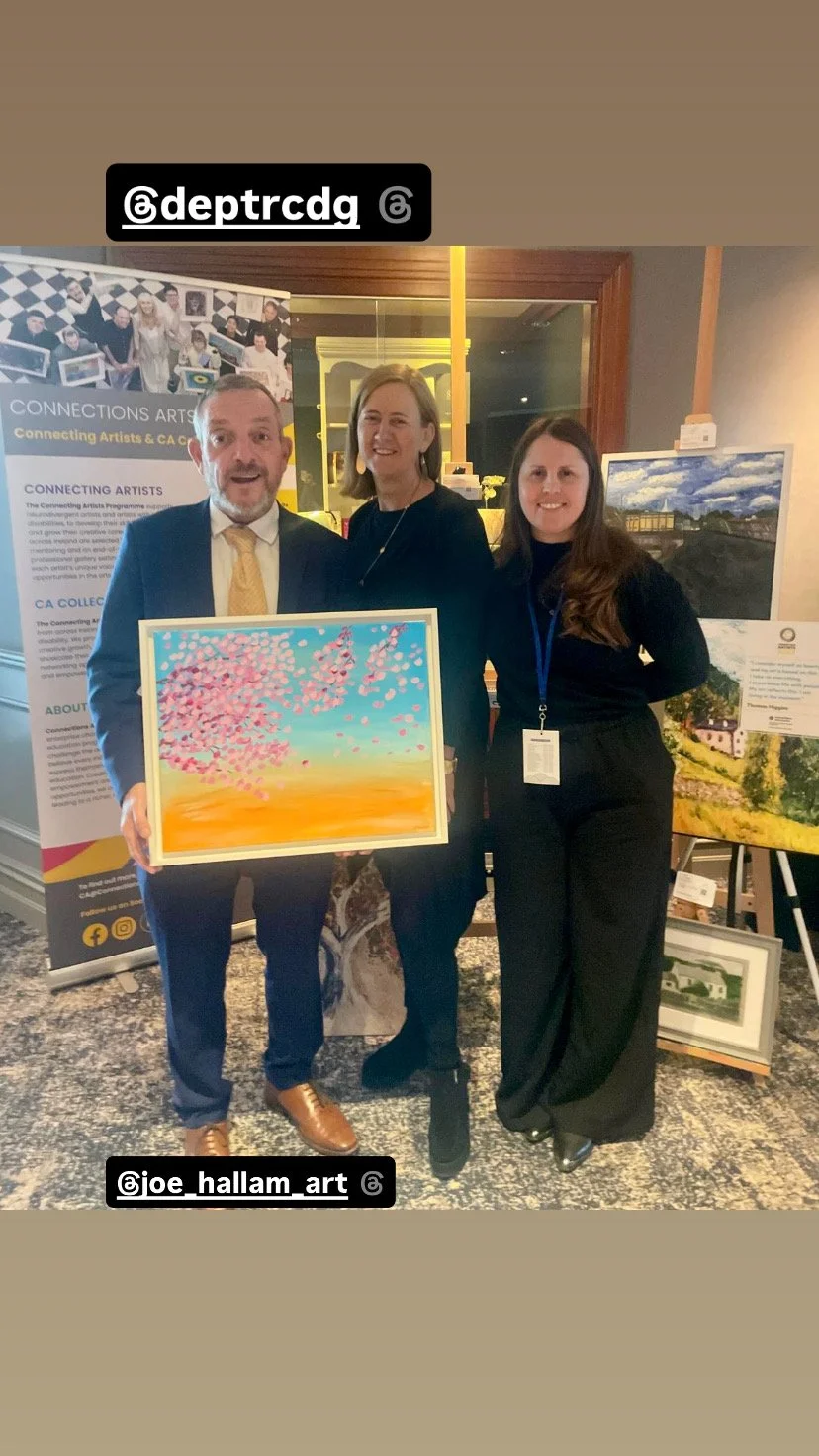 Three people standing in a group. One person is holding a painting with of a landscape, with a tree with pink flowers, blue sky, and orange ground. 