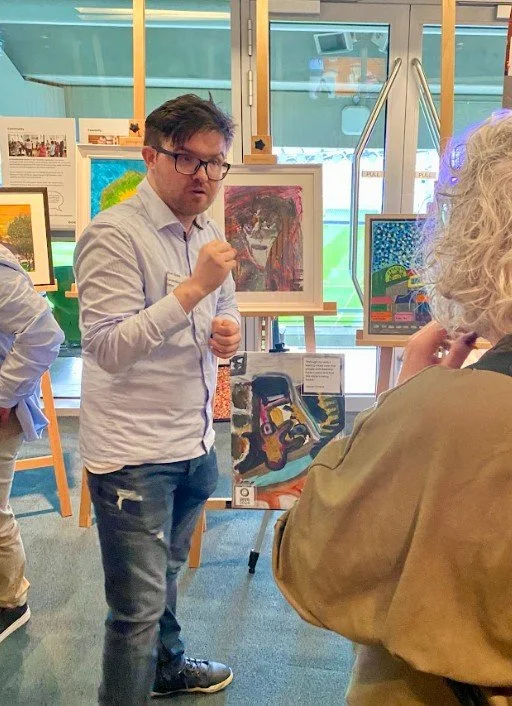 Male in a button down shirt standing in front of artwork displayed on easels. Speaking to a person who is facing them.