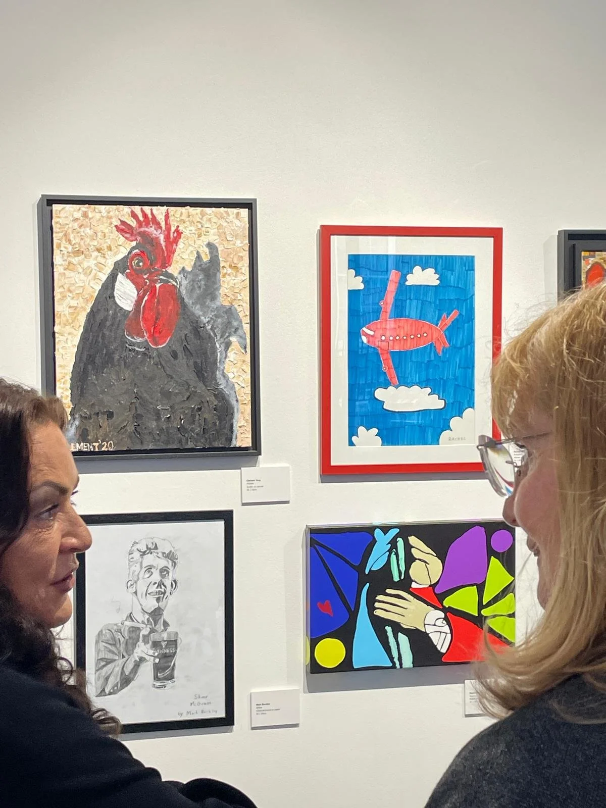 Two individuals observe framed artwork on a white wall, including a painting of a black chicken wearing a mask, a drawing of a red airplane against a blue sky, a sketch of a man holding a drink, and an abstract piece with vibrant geometric shapes.