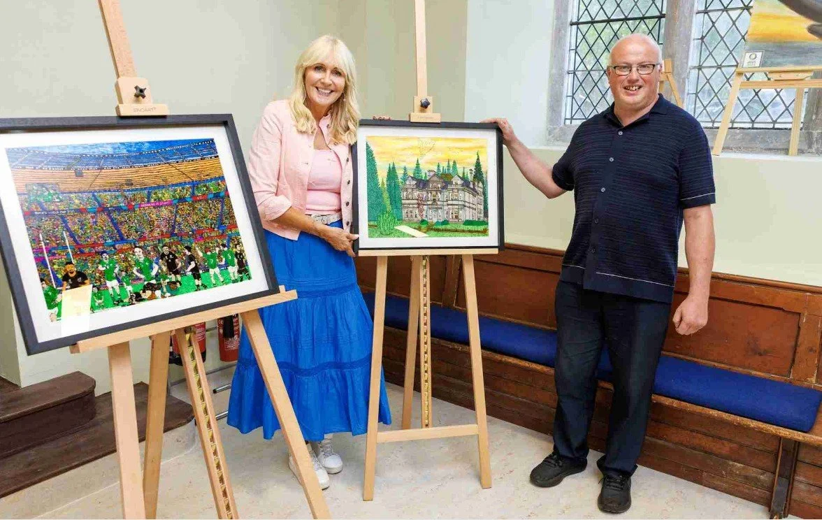 A man and a woman stand next to two framed artworks displayed on easels in a room with wooden benches and stained-glass windows. The woman, wearing a pink jacket and a blue skirt, holds a framed illustration of a crowded football stadium with players