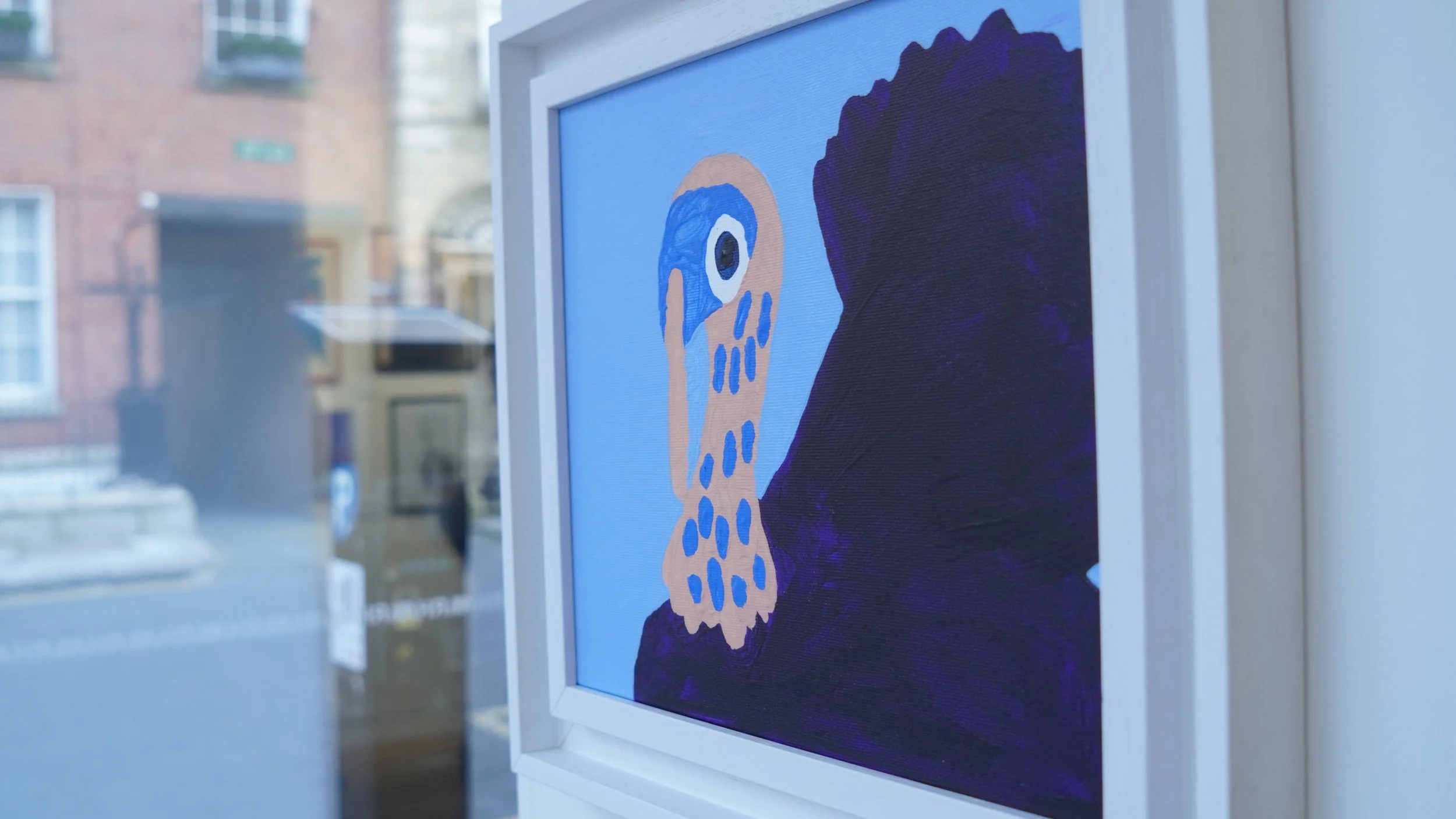 A framed, abstract acrylic painting on a light blue background. The painting features a stylized, pale orange figure with a large blue eye and blue spots, set against a dark purple, abstract shape. The painting is displayed in a white frame in a gall