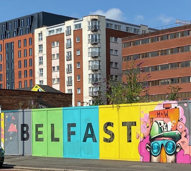 A sign painted on the side of a fence that says "BELFAST" it is multi-colored and had a graffiti style man in a hat to the right of the writing.