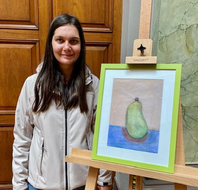 Woman stands beside a framed pastel drawing of a green pear on a blue surface, displayed on an easel.
