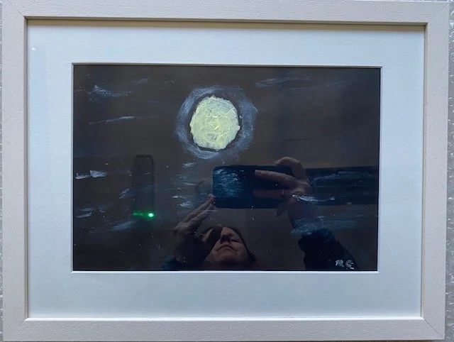 Framed abstract painting of a yellow moon in a dark sky with white brushstrokes, reflected in a mirror. The reflection also shows a person holding a phone, a green light, and the signature "RE".