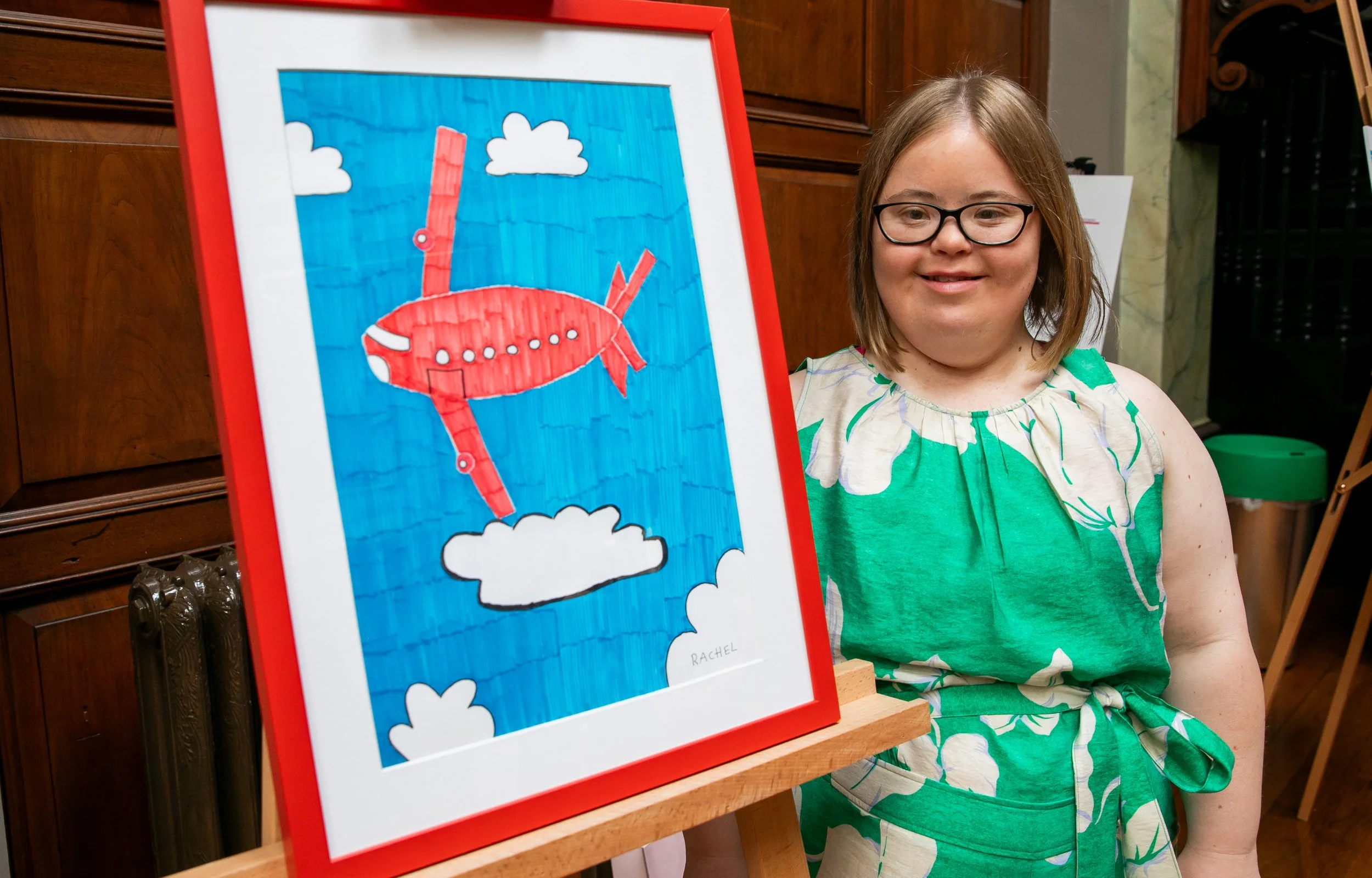 A woman wearing glasses and a green floral dress, smiling and standing beside a red-framed drawing of a red airplane in a blue sky with white clouds. The artwork, signed "Rachel," is on a wooden easel.