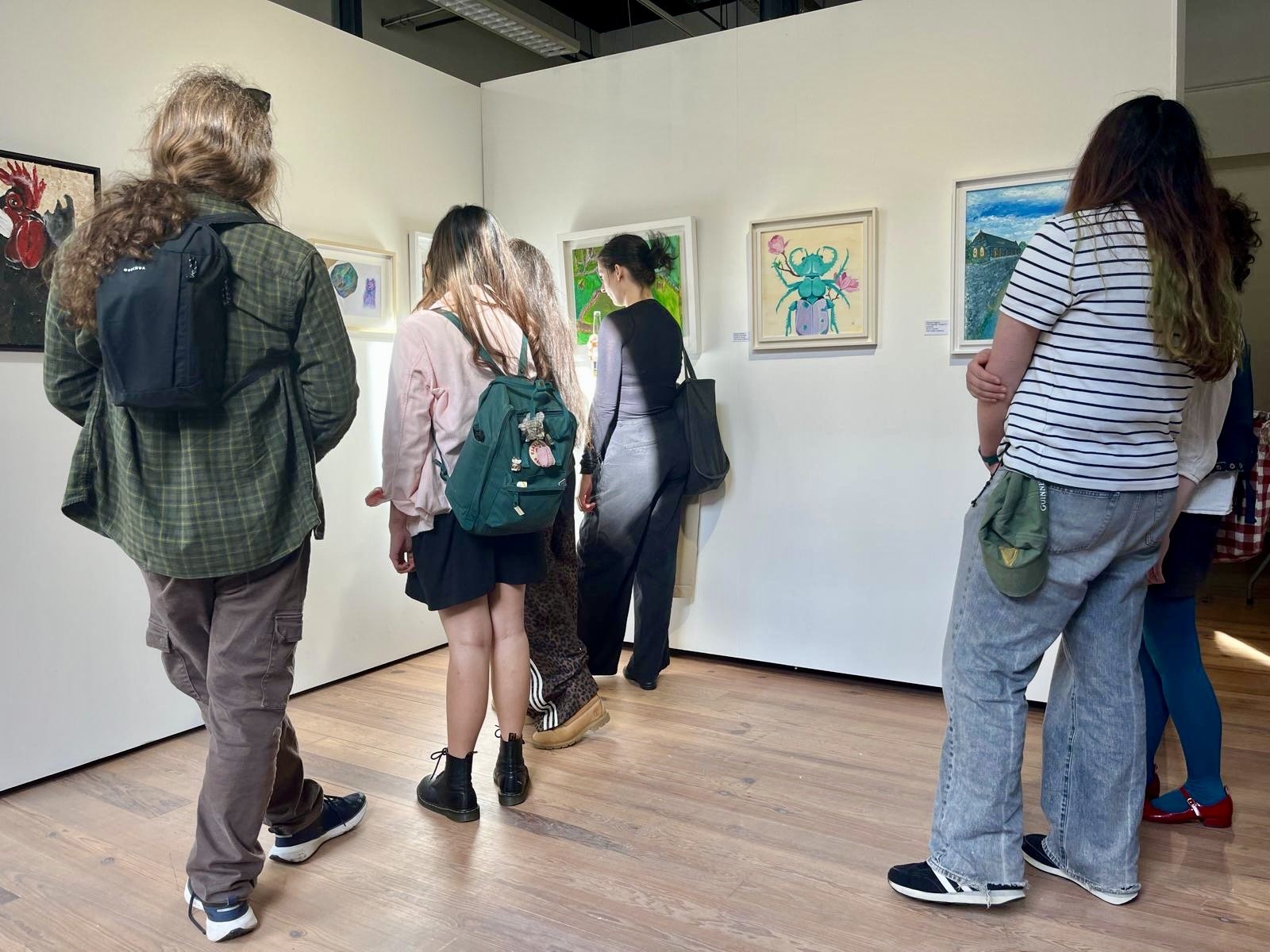 People viewing artwork on a white gallery wall, including a painting of a stylized turquoise beetle with pink flowers.