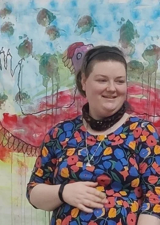A person in a short-sleeved, knee-length dress with a vibrant floral print of blue, red, and yellow flowers on a black background, stands in front of a colorful, abstract painting.