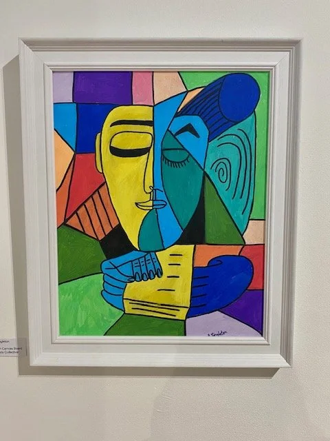 A Cubist-style painting in a white frame depicts a person with a yellow face on the left and a teal face on the right, divided vertically. The figure holds a yellow book with blue hands, and the background is a mosaic of colorful geometric shapes.