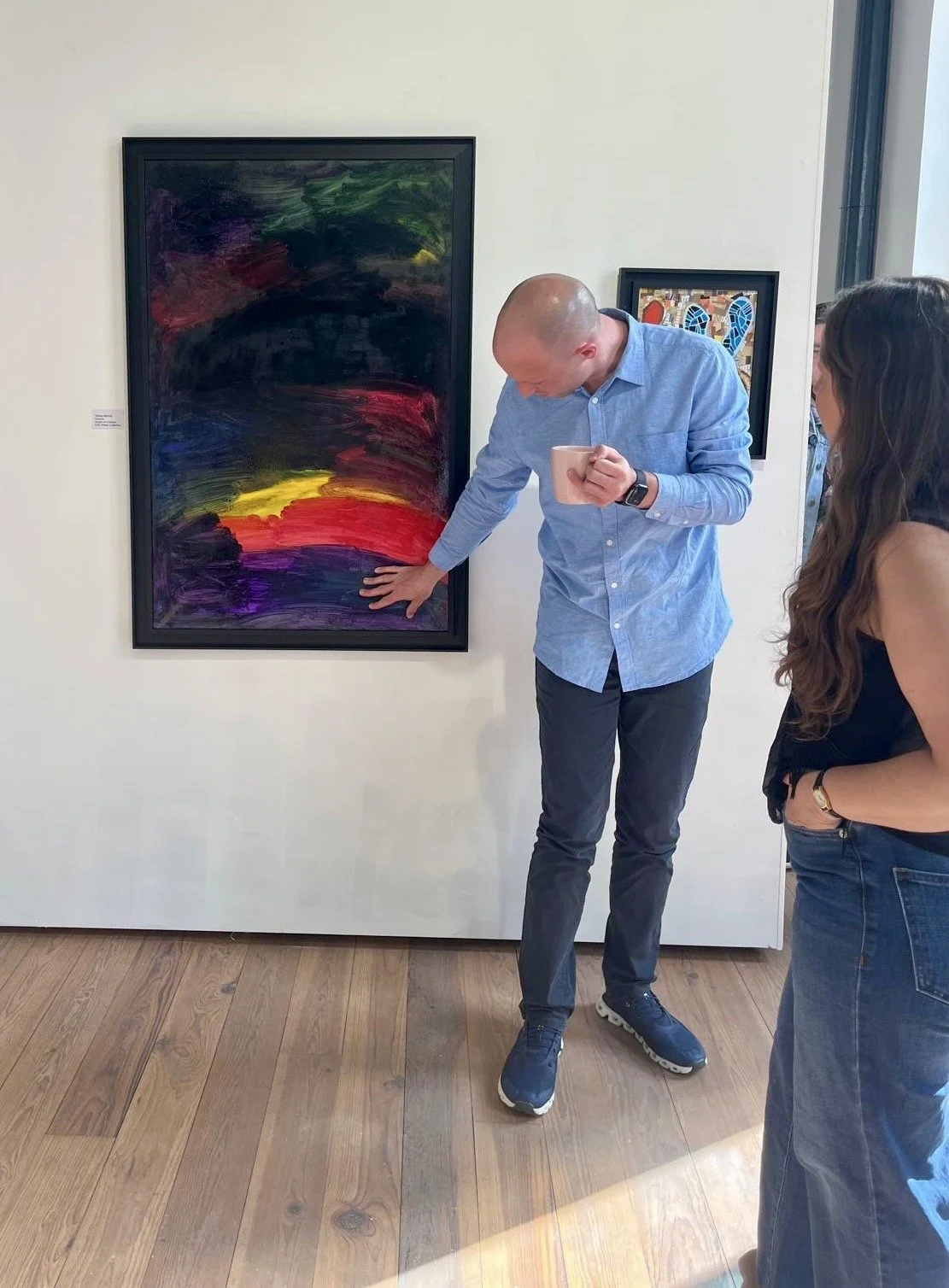 A man in a blue button-down shirt and dark pants gestures towards a large, abstract painting with bold brushstrokes of dark colors, purple, red, and yellow, while holding a mug. A woman in a black top and jeans stands to his right, looking at the art
