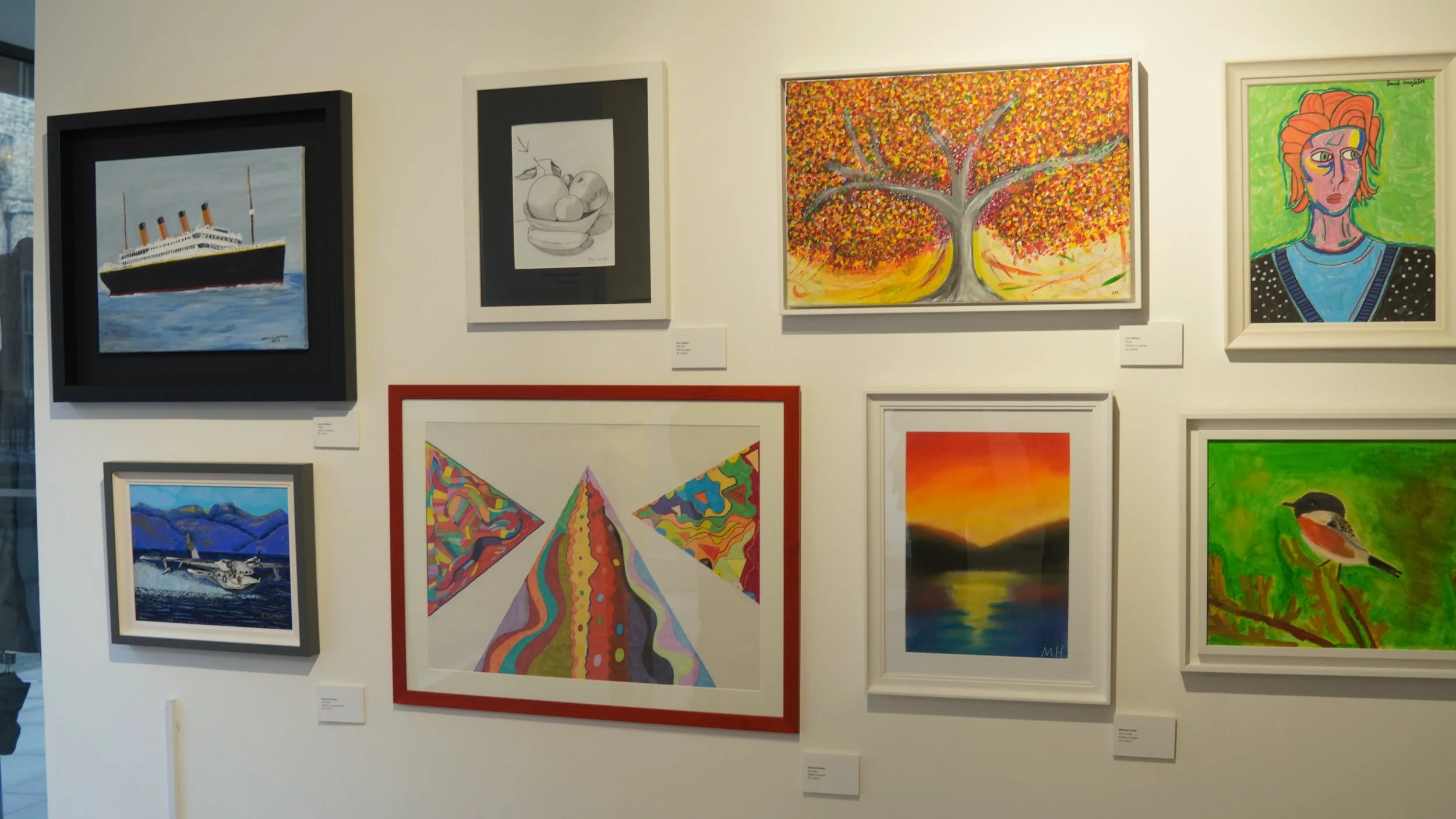 A gallery wall displays a collection of framed artworks including a painting of the Titanic, a sketch of fruit in a bowl, a vibrantly colored abstract tree, a stylized portrait, a landscape painting of a sunset over water, a painting of a bird on a b