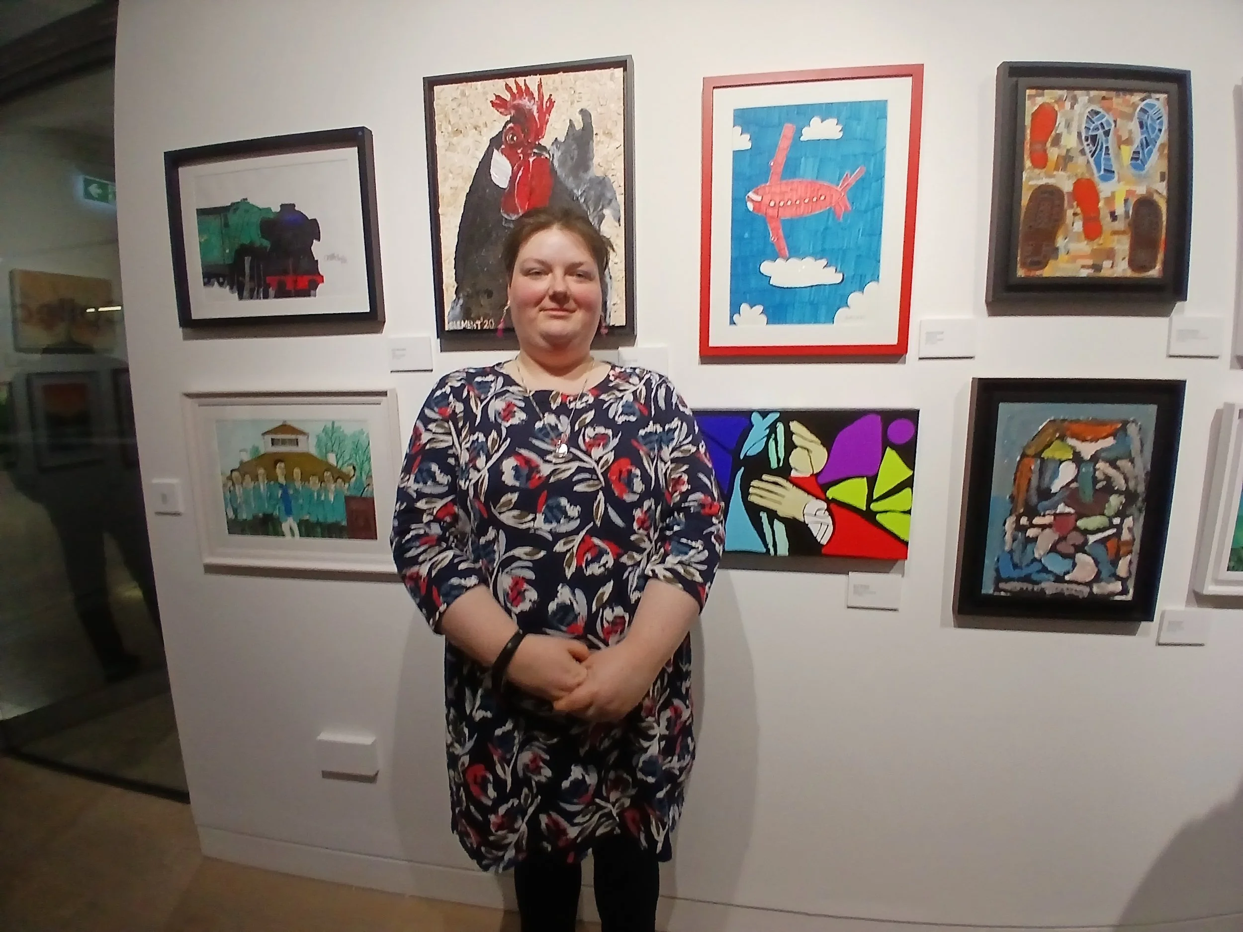 A person stands in front of a gallery wall displaying several colorful, diverse artworks, including a painting of a rooster, a stylized airplane, and a composition of abstract shapes.