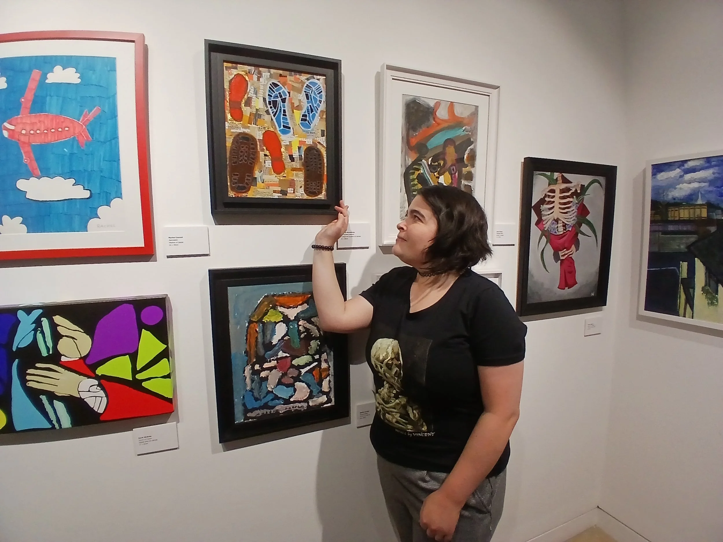 A person stands in an art gallery, looking at a collection of paintings and artwork on a white wall.