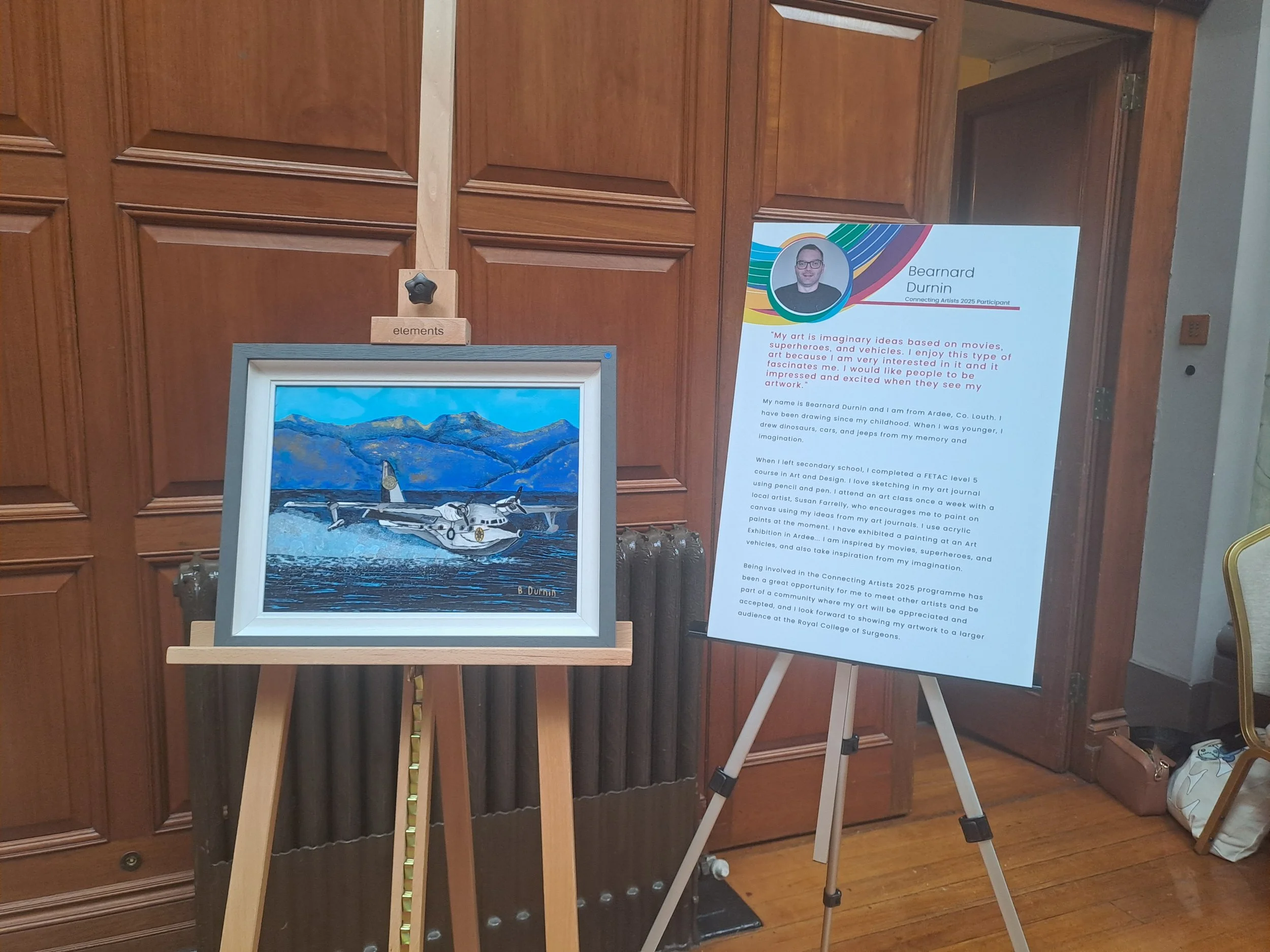 Painting on an easel depicts a white seaplane taking off from choppy water, with blue mountains in the background. To the right, a display board contains text and a portrait of an artist.