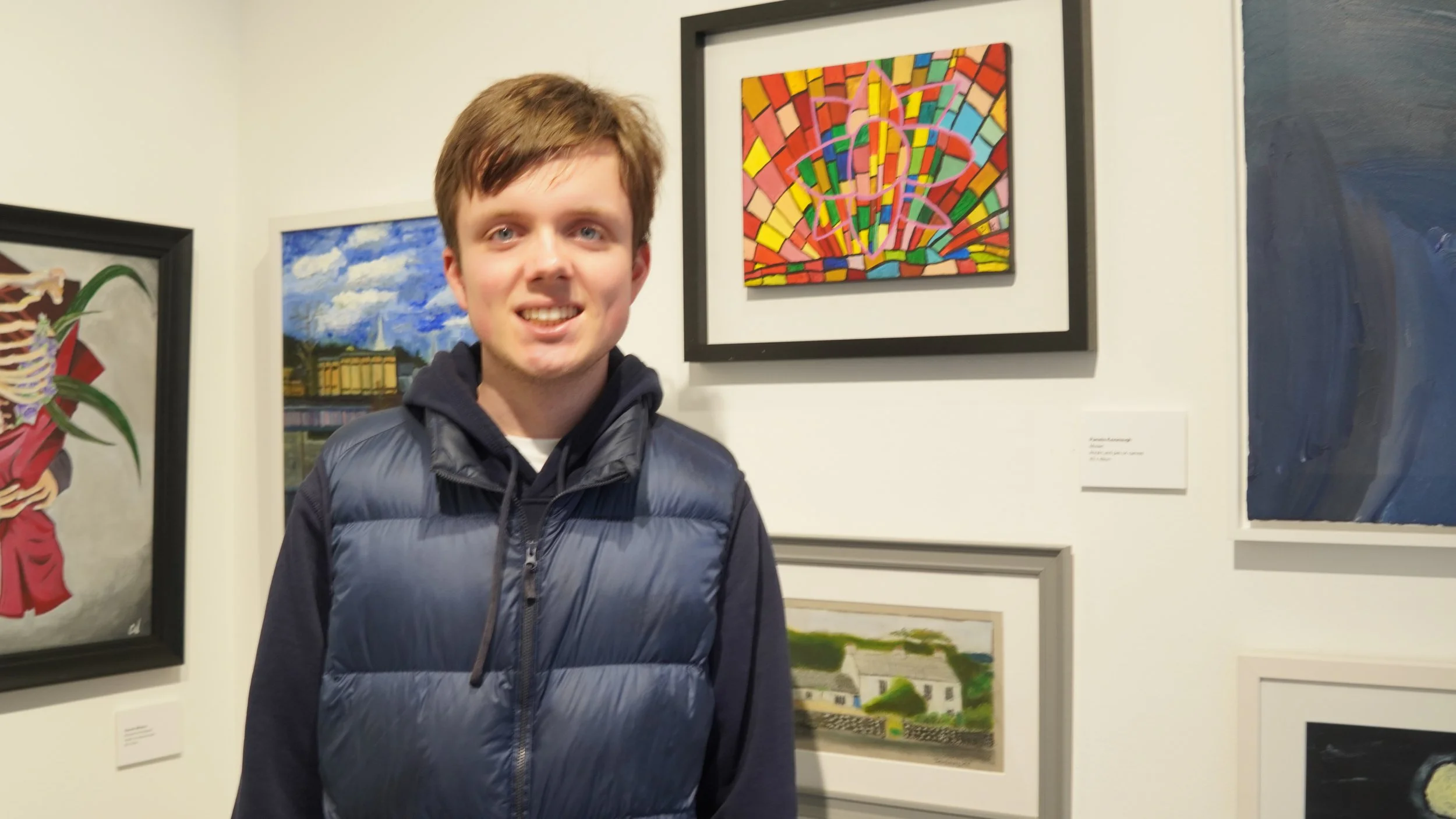 A young man wearing a blue puffer vest over a hoodie smiles at the camera in front of several framed artworks in a gallery setting.