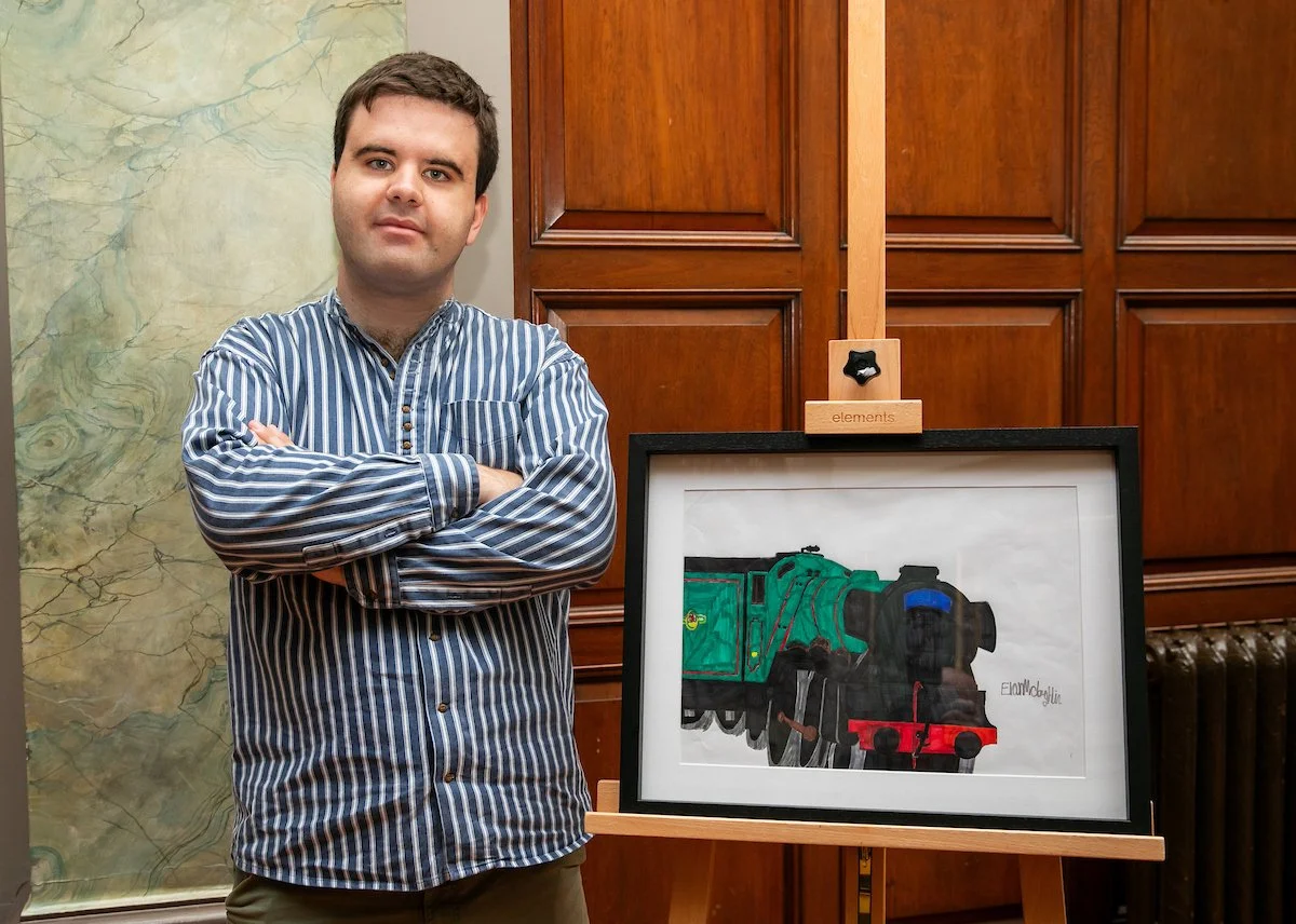 A man in a blue and white striped shirt stands with his arms crossed next to a framed drawing of a green steam locomotive on an easel.