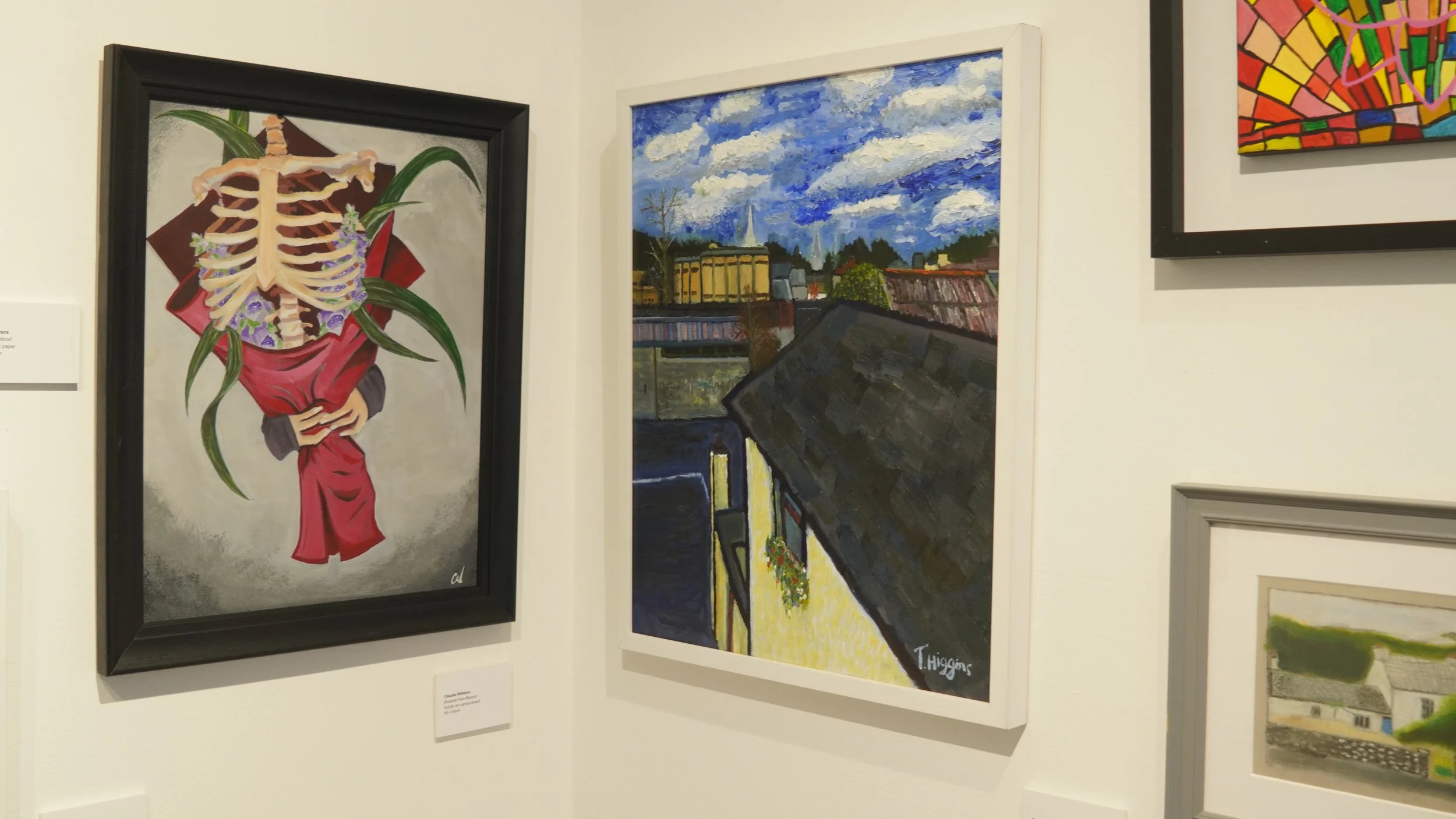 Two framed paintings displayed on a white wall: one on the left shows a human ribcage and spine intertwined with a bouquet of red fabric and purple flowers, against a textured grey background; the other painting on the right depicts a cityscape with 