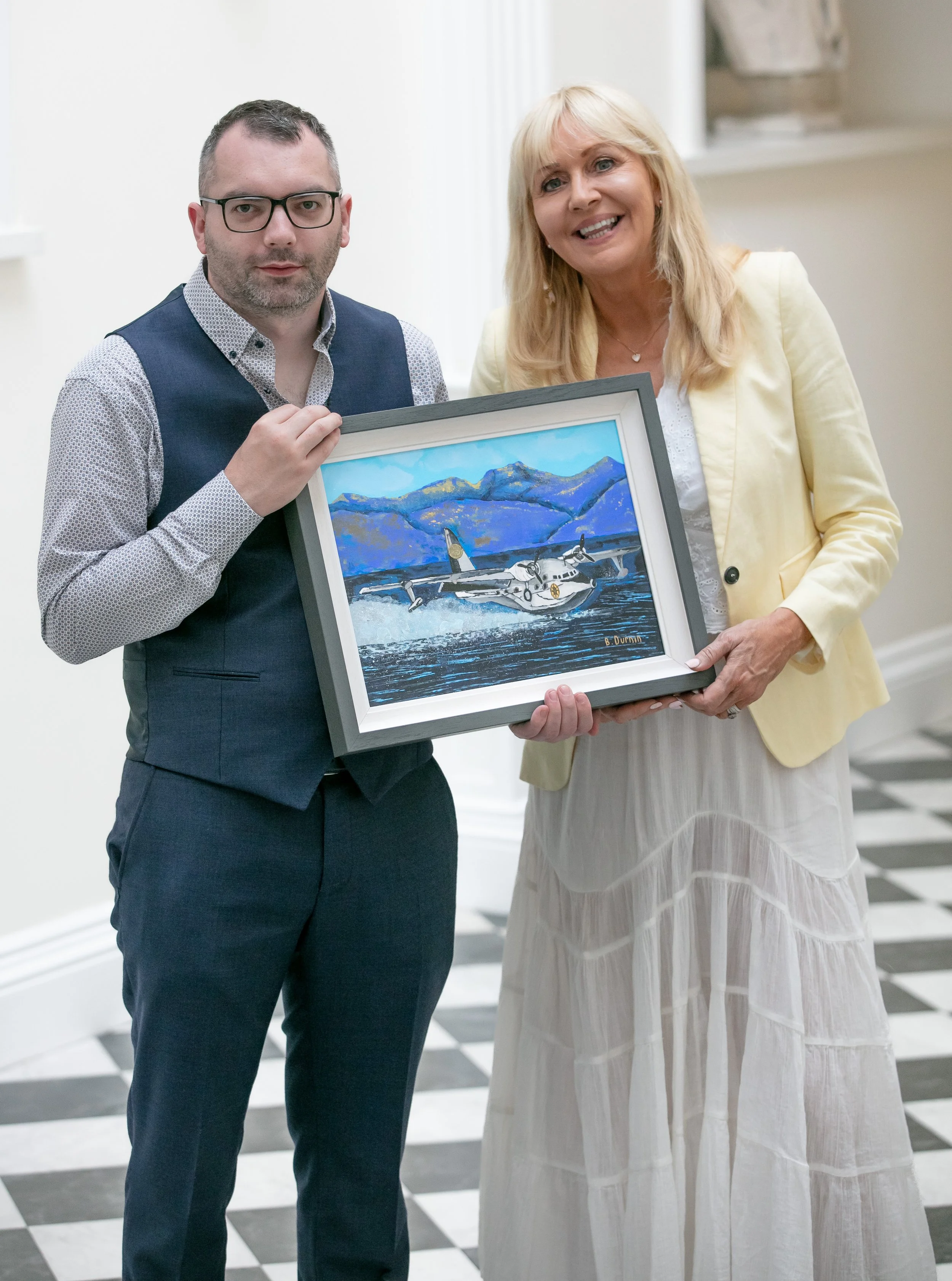 A man and a woman hold a framed painting of a seaplane on water with mountains in the background.