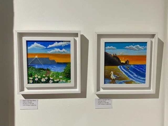 Two acrylic paintings are displayed side-by-side on a white wall. The left painting depicts a coastal landscape with a body of water, cliffs, a field of daisies in the foreground, and a bright orange and blue sky. The right painting shows a beach sce