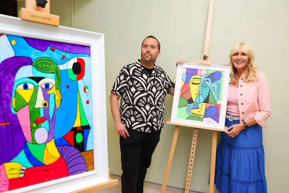 Two people stand near colorful, abstract cubist-style paintings. A man in a black and white patterned shirt stands to the left of a painting on an easel, while a woman in a pink jacket and blue skirt stands to the right of the same painting. Another
