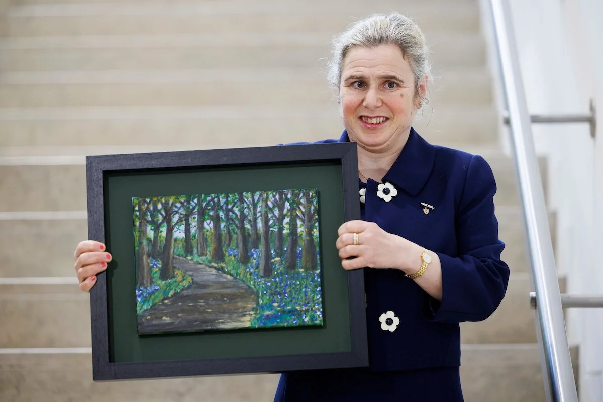 A woman in a dark blue suit holds up a framed painting of a forest path lined with trees and blue flowers.