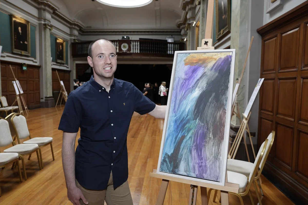 A man in a navy blue collared shirt stands next to an abstract painting of purple, blue, and gray brushstrokes on an easel in a large hall.
