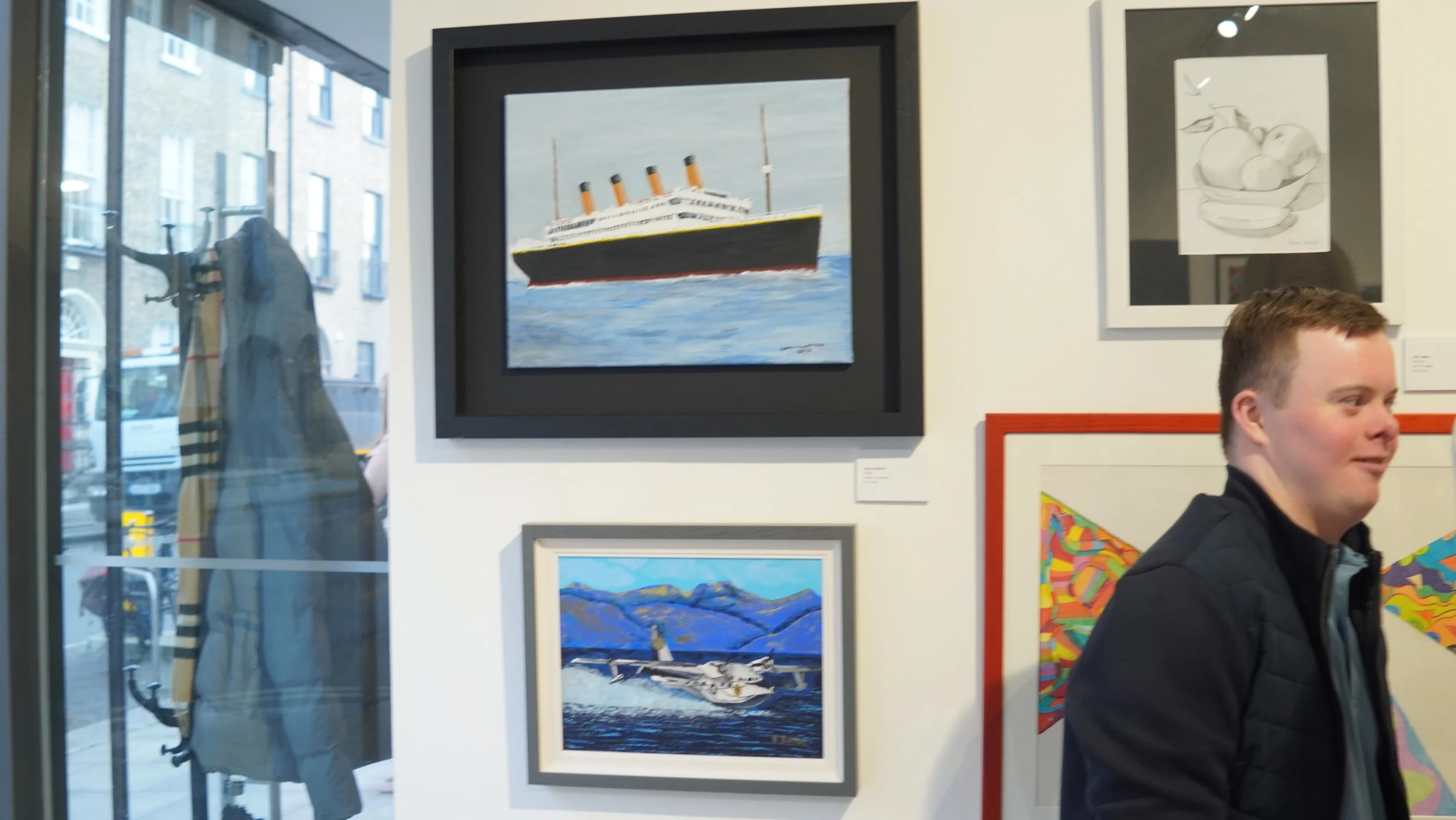 Art exhibition with paintings of a large ship and a seaplane, with a person viewing the artwork.