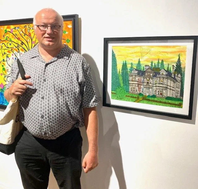 A person stands next to a framed watercolor painting of a large, ornate house surrounded by trees and a vibrant yellow sky.