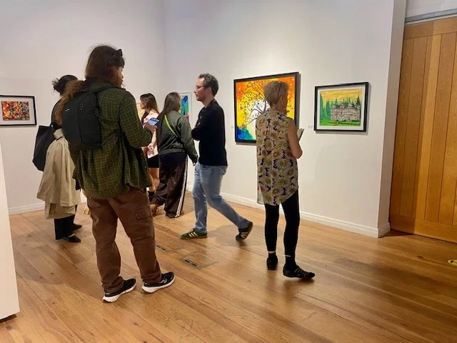 Several people are standing on a wooden floor looking at framed artwork displayed on a white gallery wall.
