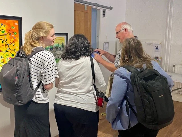 Group of people looking at art on a wall in a gallery, one person is showing something on a phone to another.