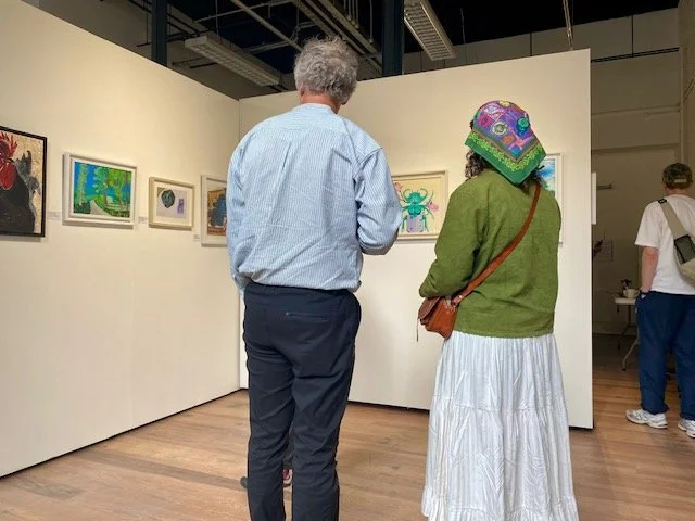 Two individuals stand in an art gallery looking at paintings displayed on white walls, including a detailed mosaic of a rooster and several abstract and nature-themed artworks.