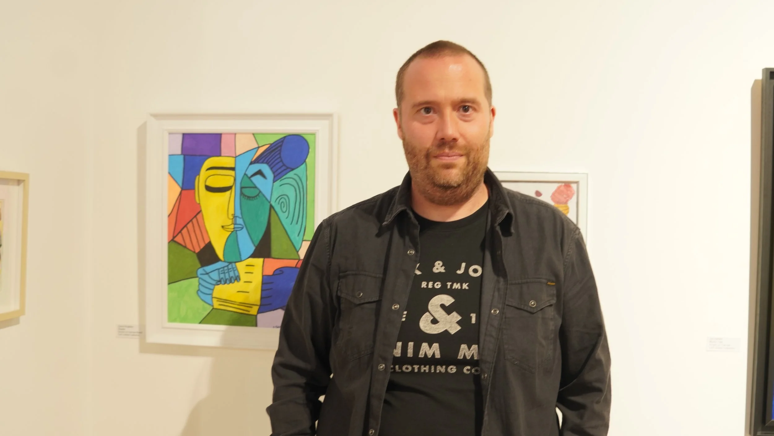A man with short brown hair and a beard stands in front of colorful abstract art displayed on a white wall in a gallery.