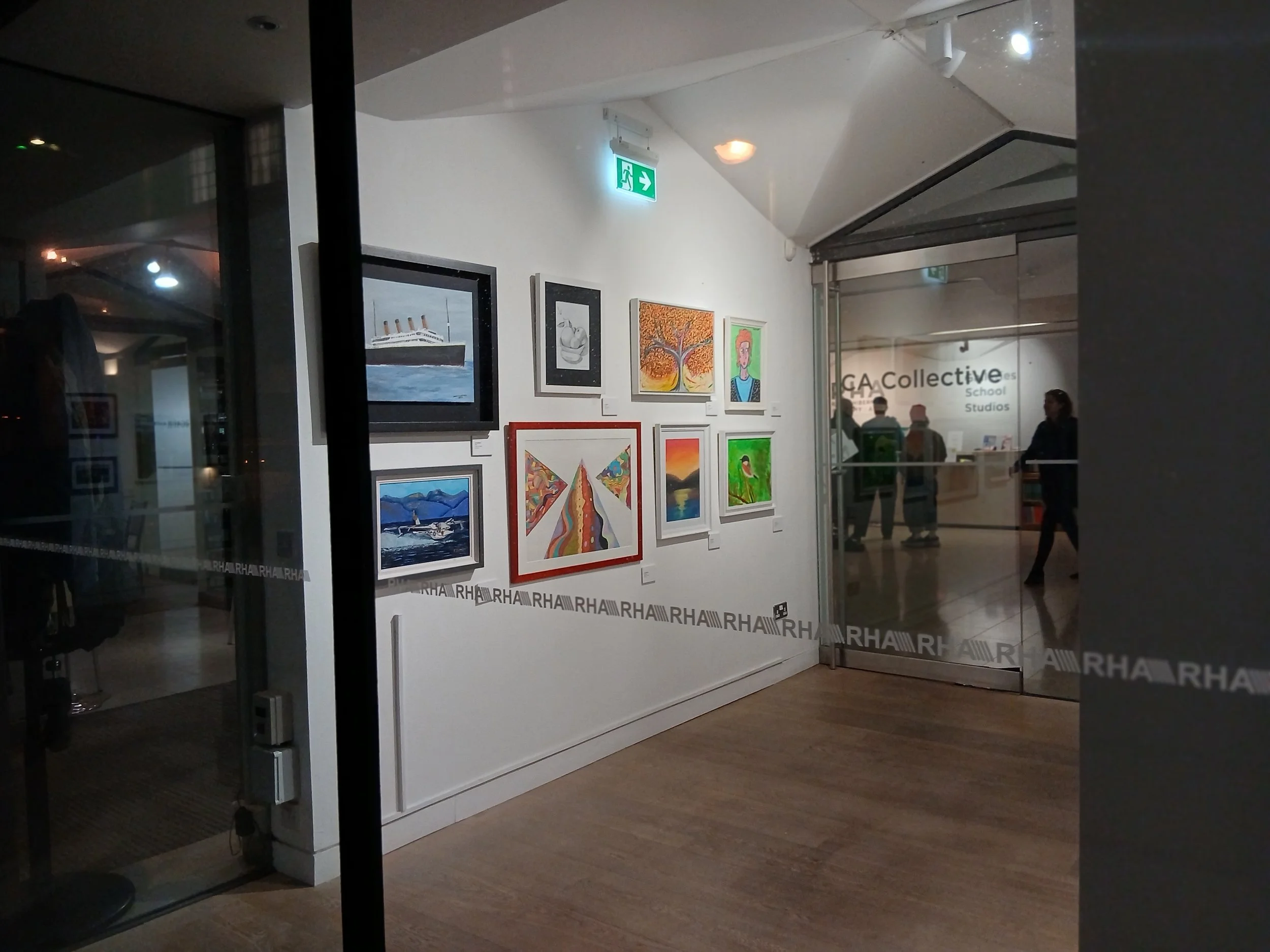 An art exhibition showcasing various paintings displayed on a white wall, with a large glass door entrance to the right leading to an interior space.